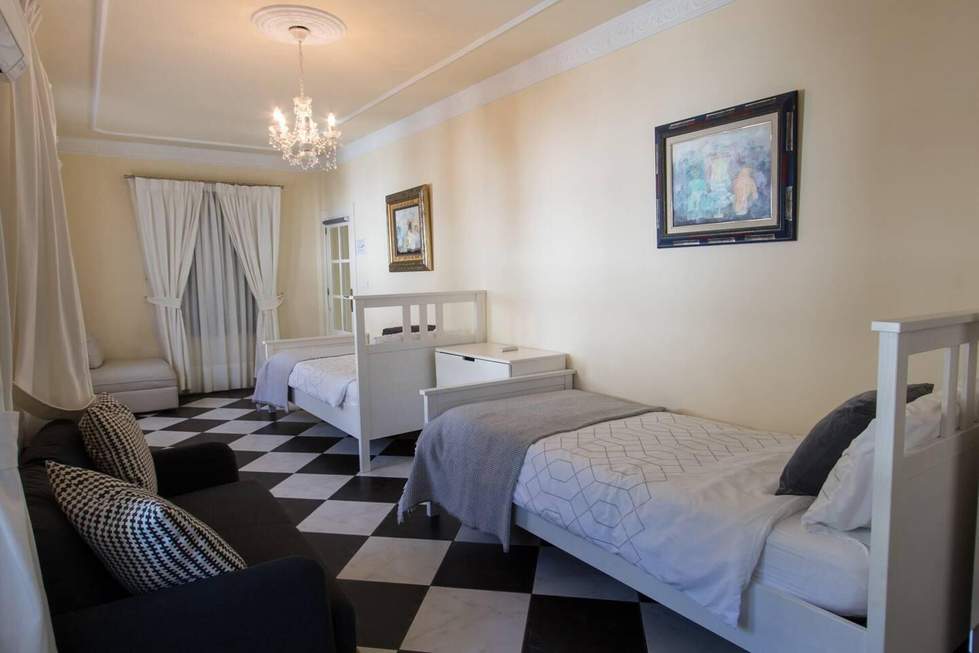 A hotel room with a checkered floor, two beds, a bedside table, a mirror, a chandelier, and a painting on the wall.