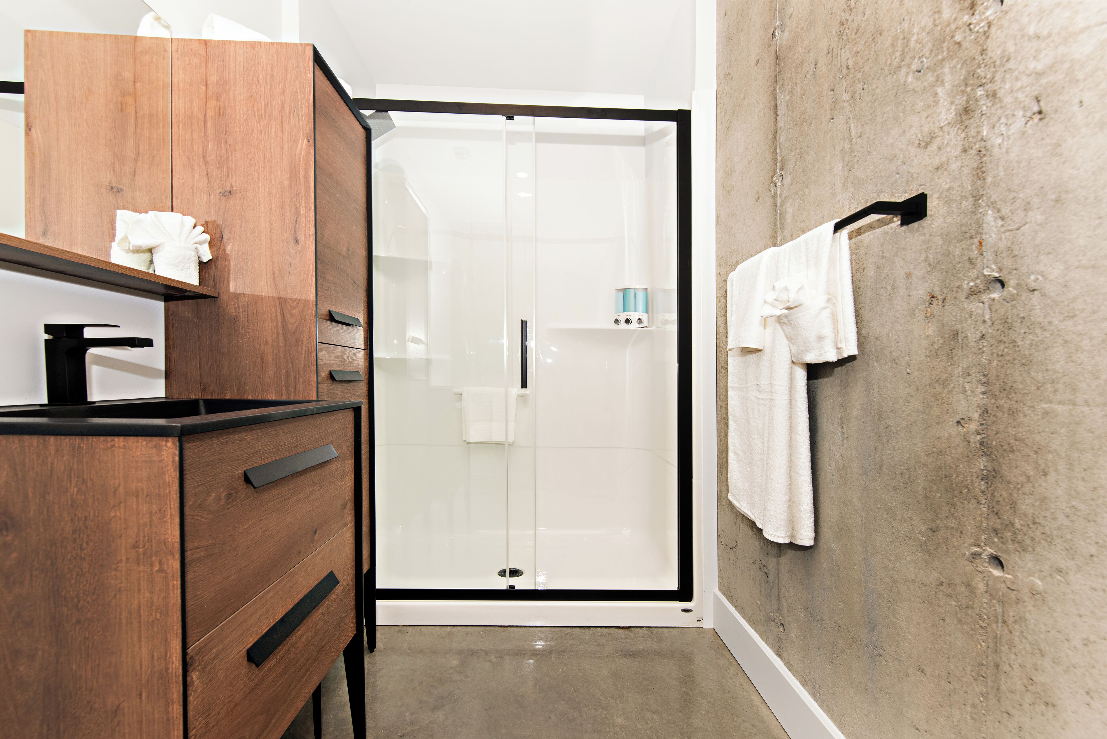 Une salle de bain moderne avec un meuble-lavabo en bois, une cabine de douche en verre et un porte-serviettes fixé à un mur en béton.