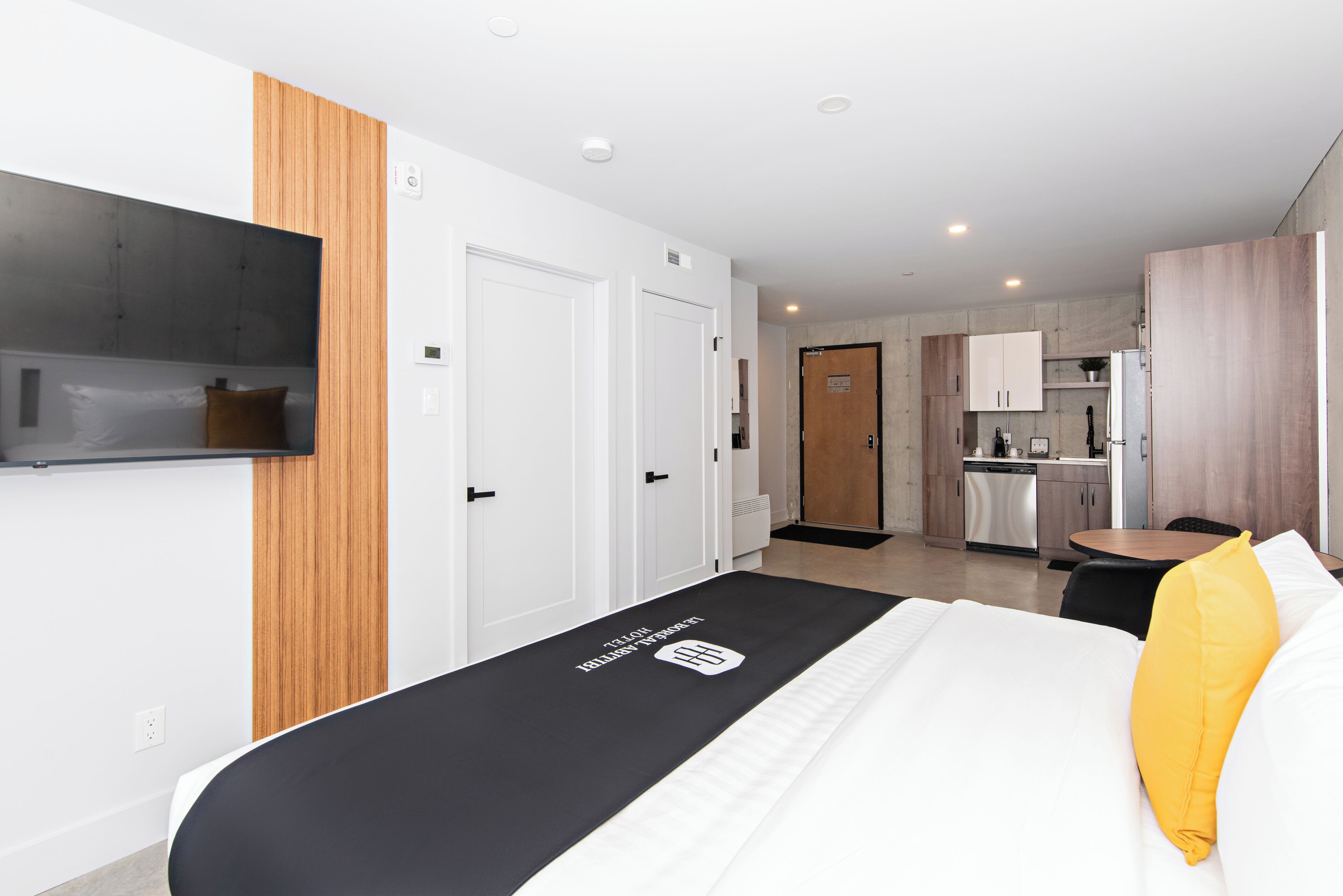 A modern hotel room with a bed, a TV mounted on the wall, a wooden panel on the wall, and a kitchen area in the background.