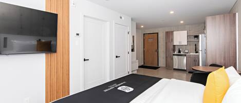 A modern hotel room with a bed, a TV mounted on the wall, a wooden panel on the wall, and a kitchen area in the background.