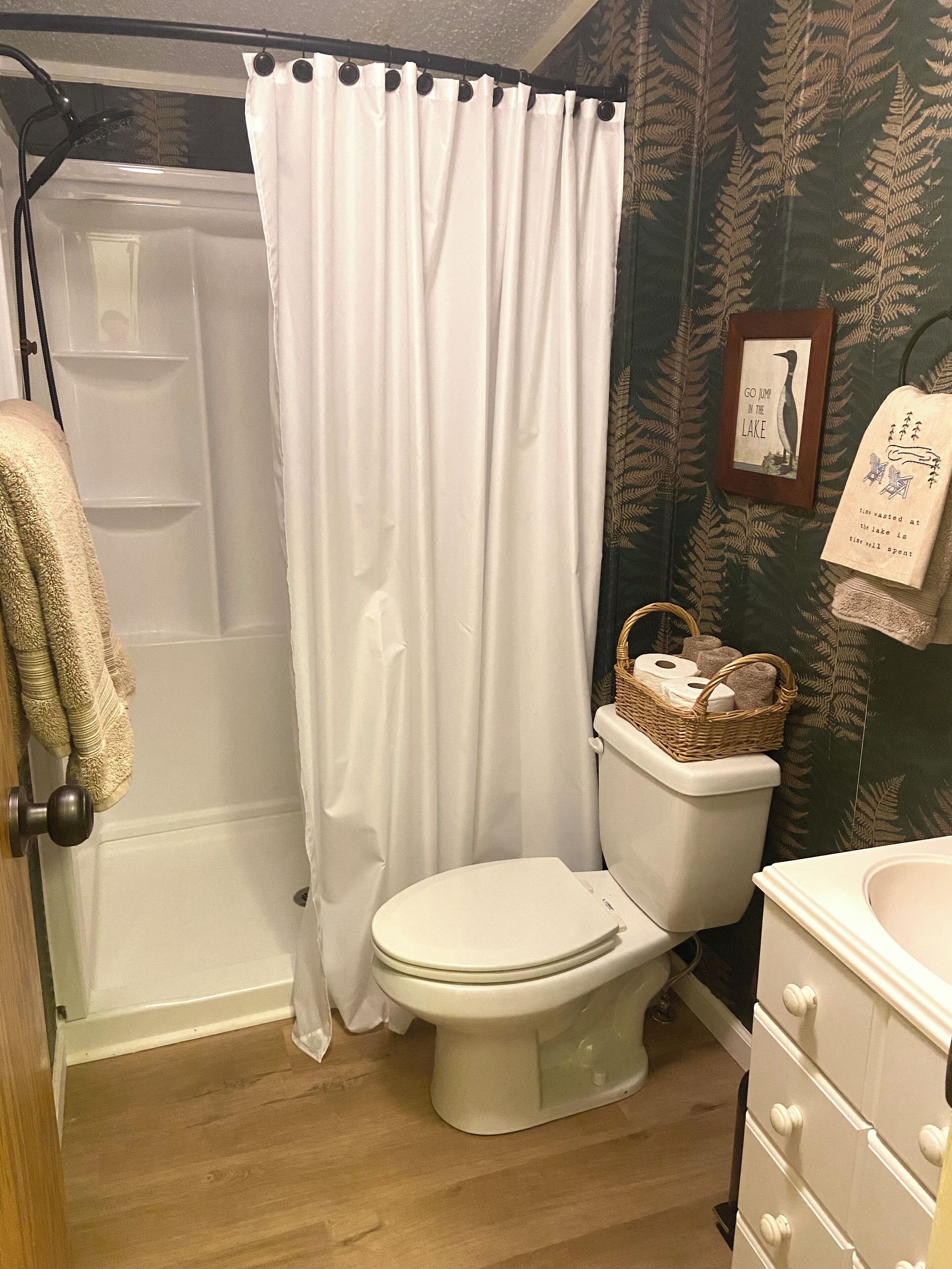 A bathroom with a shower curtain, a toilet, a wicker basket with toiletries, and a framed picture on the wall.