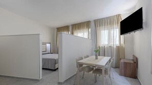Ein modernes Hotelzimmer mit einem Bett, einem Esstisch mit Stühlen, einem an der Wand befestigten Fernseher und einem kleinen Holzhochentisch.