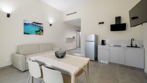 Eine moderne Küche mit Einbauofen, Kühlschrank und Kaffeemaschine. Es gibt eine Essecke mit Tisch und Stühlen sowie eine Couch. An der Wand hängt ein Gemälde, das eine Küstenlandschaft zeigt.