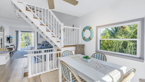 A bright, airy living room with a white staircase, a dining area with a white table and blue chairs, and a large window with a view of palm trees. - Reese's Beach Retreat on beautiful Anna Maria Island (Holmes Beach)
