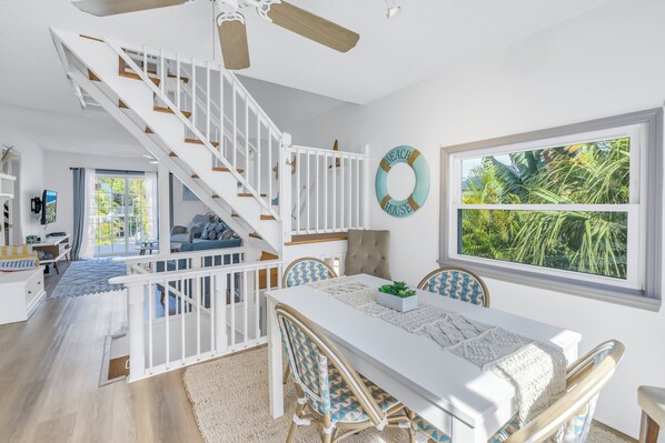 A bright, airy living room with a white staircase, a dining area with a white table and blue chairs, and a large window with a view of palm trees. - Reese's Beach Retreat on beautiful Anna Maria Island (Holmes Beach)