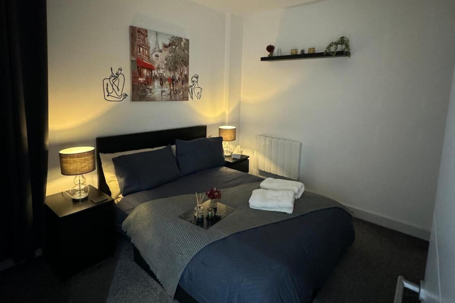 Een keurig opgeruimde hotelkamer met een groot bed, twee nachtkastjes met lampen, een radiator en een wandplank met decoratieve objecten.