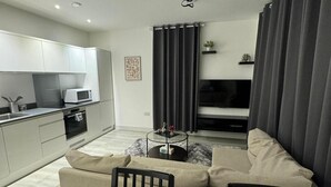 A modern living room with a sofa, a coffee table, and a TV. There is a kitchen area with cabinets and a microwave.