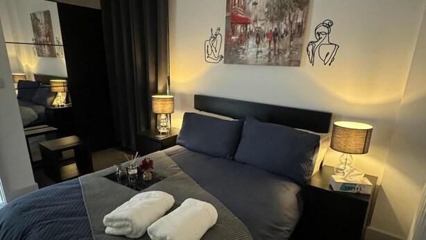 A modern hotel room with a large bed, two bedside tables with lamps, a small table with a vase and candles, and artwork on the walls.