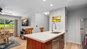 A modern kitchen with a large island, wooden cabinets, a stainless steel refrigerator, and a dining area with a view of the outdoors.