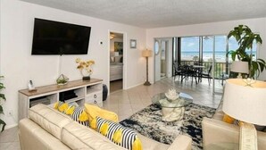 A modern living room with a flat-screen TV, a sofa with yellow and black cushions, a coffee table with decorative items, and a balcony with a view of the ocean. - Gulf Views 2 Bed 2 Bath at Siesta Key Beach 100 Steps to the Sand (Siesta Key)