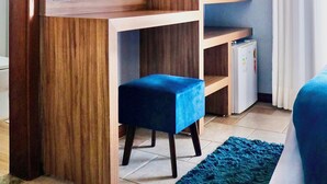 A compact room with a wooden desk, a television mounted on the wall, a small refrigerator, and a blue chair. - Lara's Hotel (Capitólio)