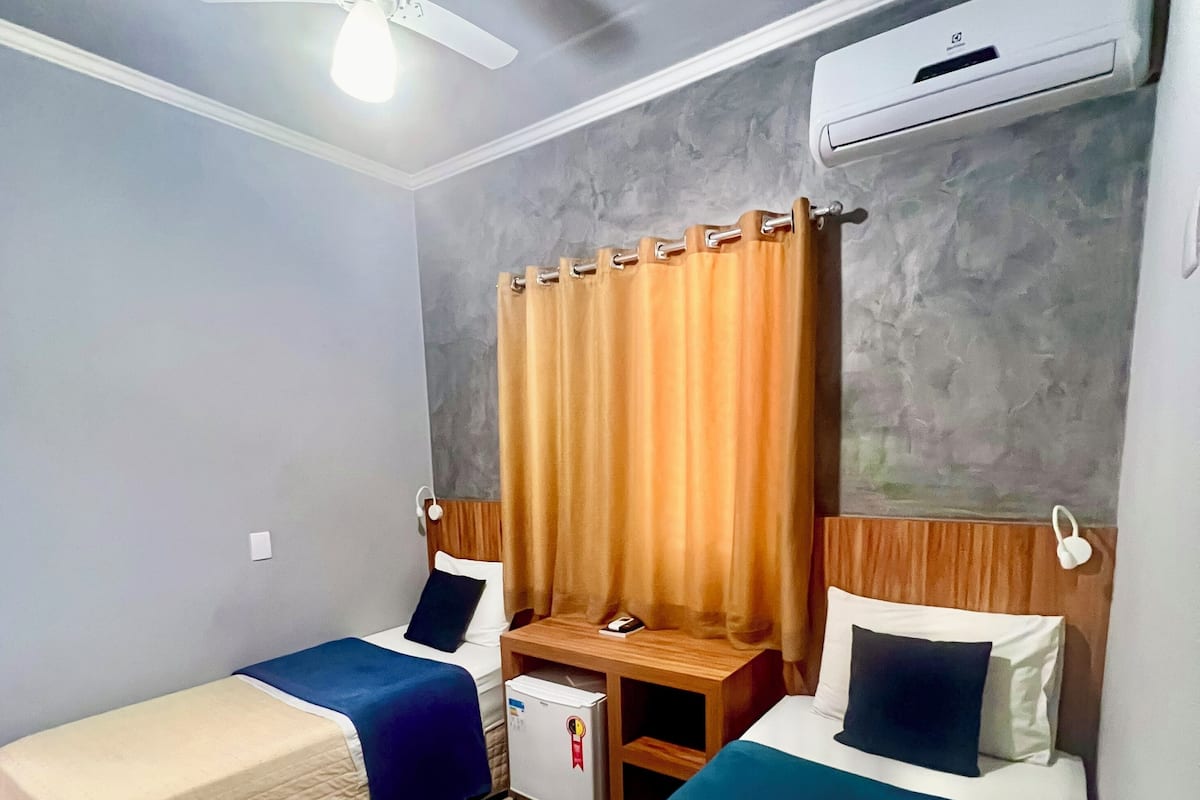 Ein Hotelzimmer mit zwei Betten, einem Deckenventilator, einer Klimaanlage und einem kleinen Kühlschrank.