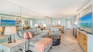 A spacious living room with a grey sofa, a coffee table, and a TV. There is a large mirror on one wall and a beach-themed decor. - Best View on Siesta Key 1 Bed 1 Bath Top Floor Steps to Beach (Siesta Key)