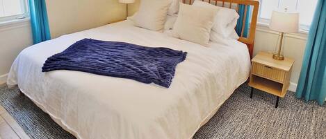 Una cama bien hecha con sábanas blancas y una manta azul, con dos lámparas de noche sobre mesitas de madera, en una habitación con paredes de colores claros y una ventana con cortinas azules.