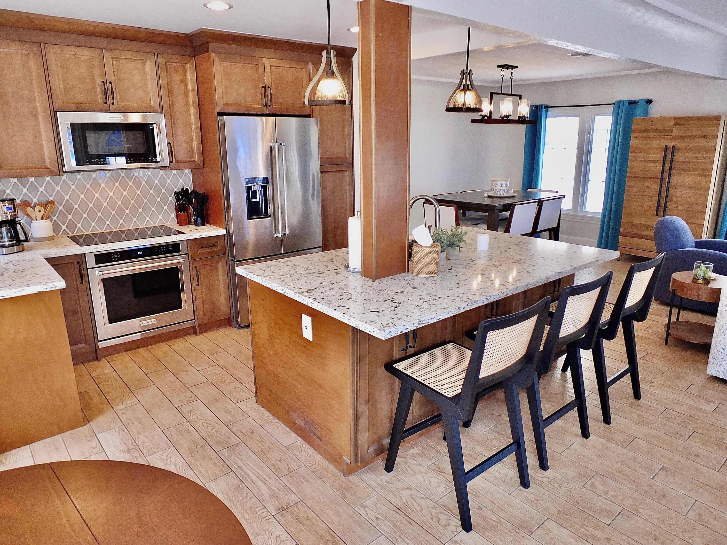A modern kitchen with stainless steel appliances, a central island with bar stools, wooden cabinets, and a dining area with blue curtains.