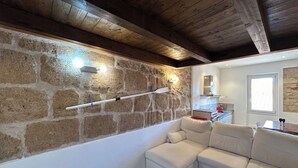 A room with a stone wall, wooden ceiling beams, a white sofa, and a kitchen in the background. - Alghero my Love (Alghero)