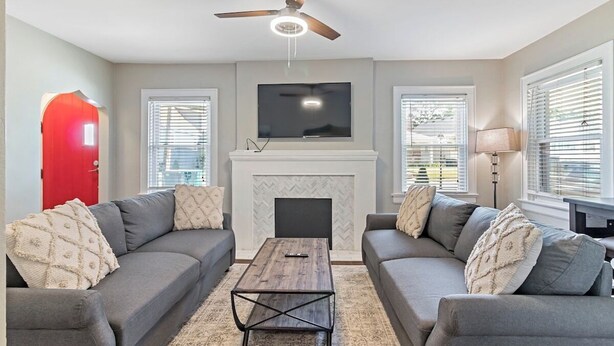 A living room with two grey sofas, a wooden coffee table, a fireplace, a ceiling fan, and a TV mounted above the fireplace.