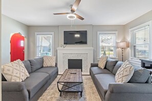 A living room with two grey sofas, a wooden coffee table, a fireplace, a ceiling fan, and a TV mounted above the fireplace.