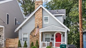 A two-story house with a gray siding exterior, a red front door, and a small porch with a white railing. There are two green trash bins and a wooden fence in the front yard.