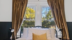 A bed with white bedding and a single gold pillow, flanked by two black nightstands, each with a lamp. The room has a window with curtains, and the walls are painted in two colors. - Peaceful Oakley Escape 2bd|Lg Priv Deck w views (Cincinnati)
