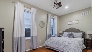 Quarto com cama, ventoinha no teto, mesa de cabeceira, abajur e janela com cortinas.