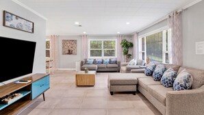 A living room with a flat-screen TV, a wooden coffee table, two sofas with patterned cushions, and a large window with blinds.