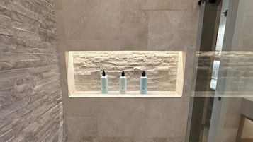Une salle de bain avec un mur en pierre, une cabine de douche en verre et une étagère sur laquelle sont posées trois bouteilles.