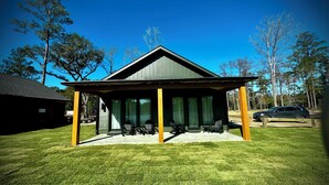 A covered outdoor seating area with chairs and tables, surrounded by a lawn and trees. - Egret Cottage Suite    3715-B (WALTERBORO)