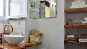 Ein Badezimmer mit einem großen weißen Waschbecken, einem Holz-Waschtisch, einem Spiegel und Regalen mit Handtüchern und Pflegeprodukten.