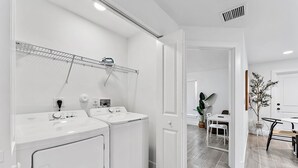 A modern laundry room with two white washing machines, a drying rack, and a small table with chairs. - Contemporary 3-Bed,2-Bath Home, Downtown WPB! (West Palm Beach)