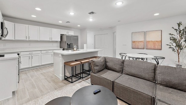 A modern kitchen with white cabinets, a central island, and a bar area. There is a gray sofa and a small dining table with chairs. - Contemporary 3-Bed,2-Bath Home, Downtown WPB! (West Palm Beach)