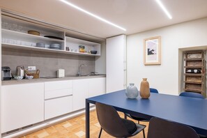 Une cuisine moderne avec des meubles blancs, une table bleue et deux chaises. On y trouve un évier, une bouilloire et divers ustensiles de cuisine sur le plan de travail.