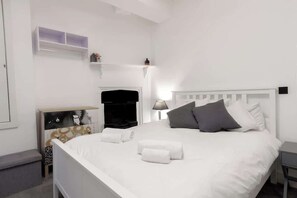 Une pièce blanche comprenant un lit, une table de chevet, une commode et une étagère fixée au mur.