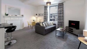 A modern living room with a sofa, a TV, a dining area with chairs, and a kitchen area with a counter and cabinets. - Chic Centrally Located 1Bed Apt in Chester (Chester)