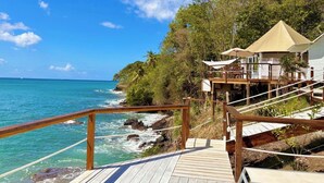 A wooden deck leading to a beachside resort with a view of the ocean and a palm tree-lined path. - Waterfront Luxury Tented Experience with Saltwater Infinity Pool & Beach - 2 Bed (Vigie)