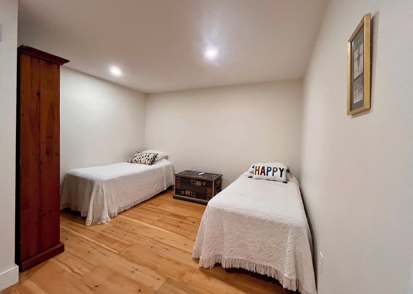 Um quarto pequeno e limpo com duas camas de solteiro, piso de madeira e uma pequena cômoda.