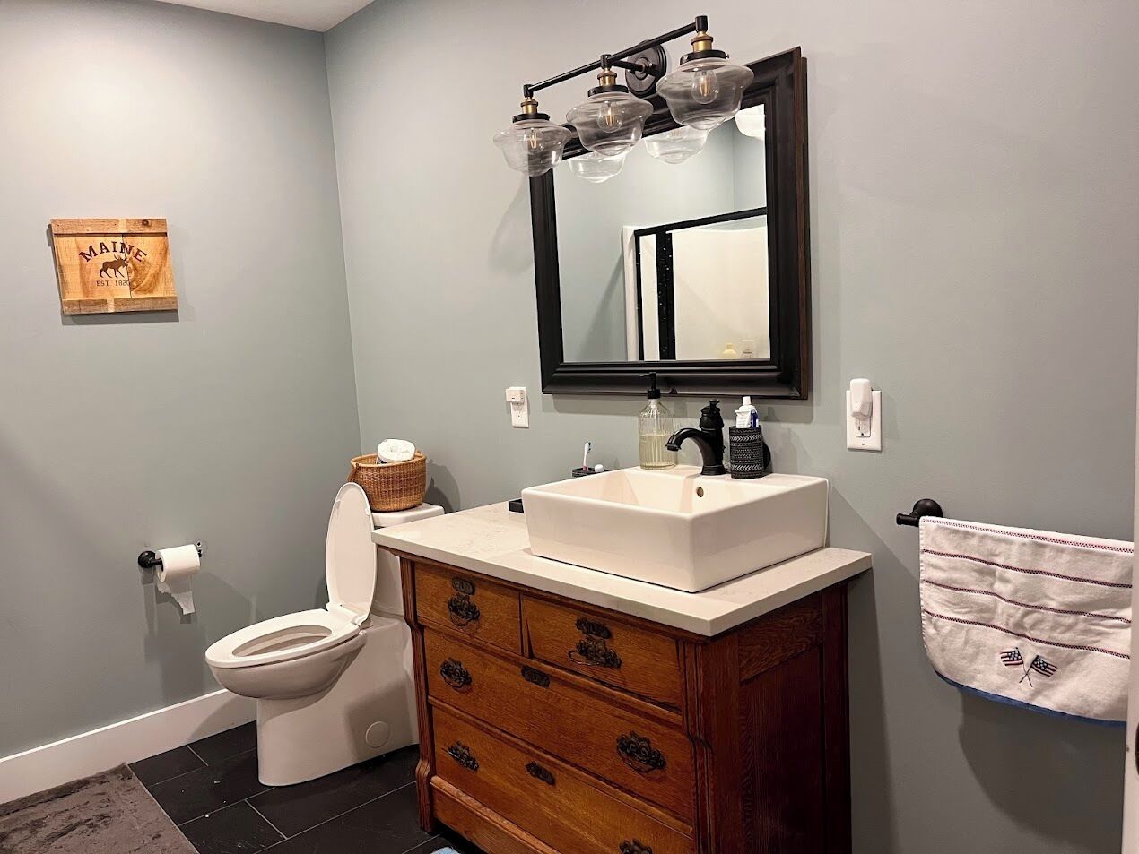 A bathroom with a toilet, a sink with a mirror above it, wooden cabinetry, and a towel hanging on the wall.