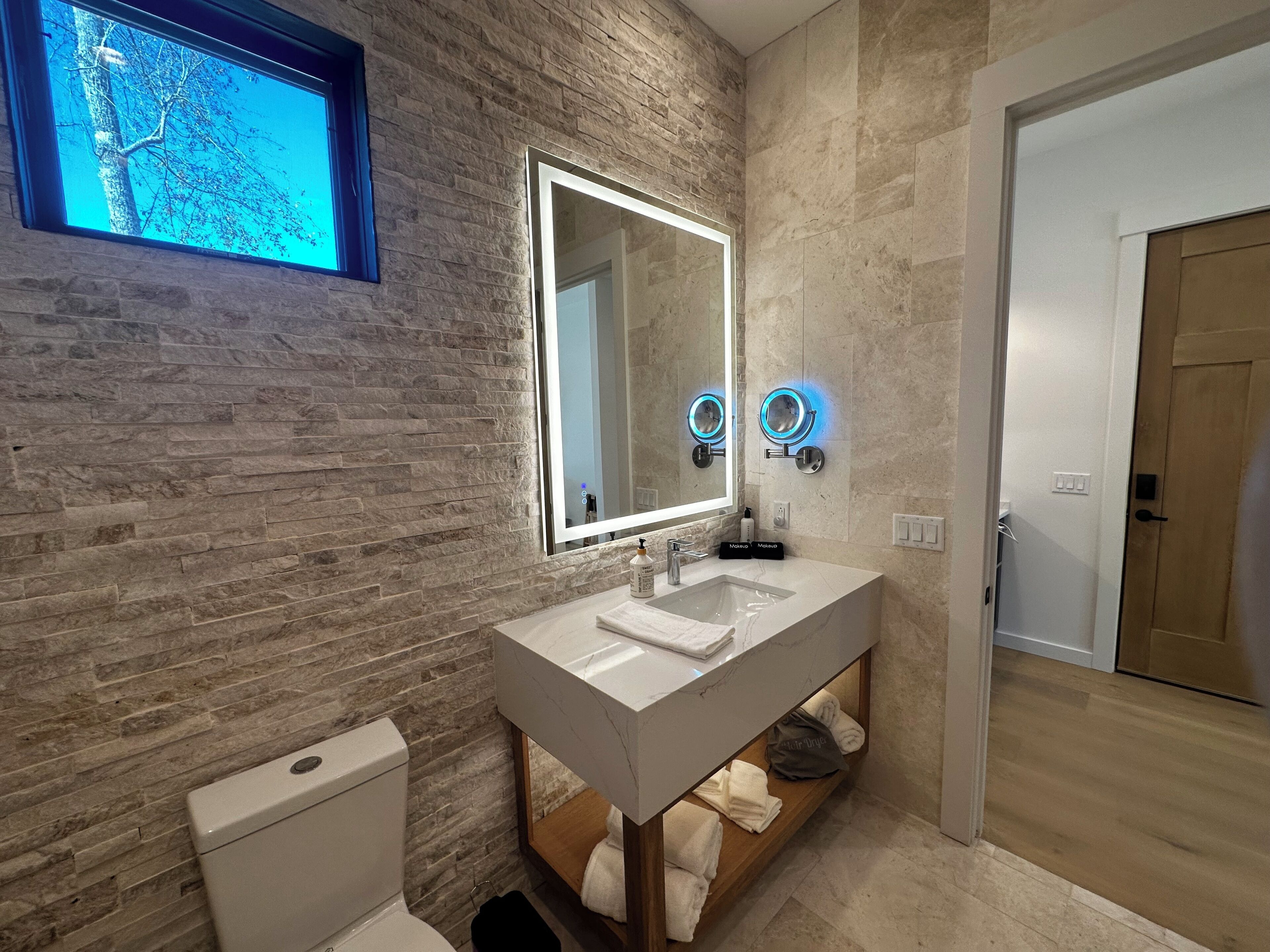 A modern bathroom with a stone wall, a large mirror, two wall-mounted lights, a white sink, and a toilet.