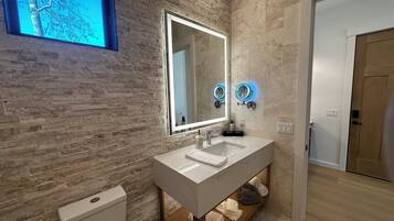 A modern bathroom with a stone wall, a large mirror, two wall-mounted lights, a white sink, and a toilet.