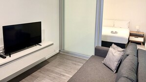 A modern hotel room with a flat-screen TV on a stand, a grey sofa with cushions, a bed with white bedding, and a nightstand with a lamp. - 2BR in Culture & Business Center (Melbourne)