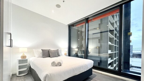 A modern bedroom with a large bed, a bedside table, a nightstand, and a large window with a view of a building. - 2BR in Culture & Business Center (Melbourne)