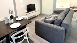 A modern living room with a grey sofa, a dining table with white chairs, a flat-screen TV mounted on the wall, and a large window providing a view of the room. - 2BR in Culture & Business Center (Melbourne)