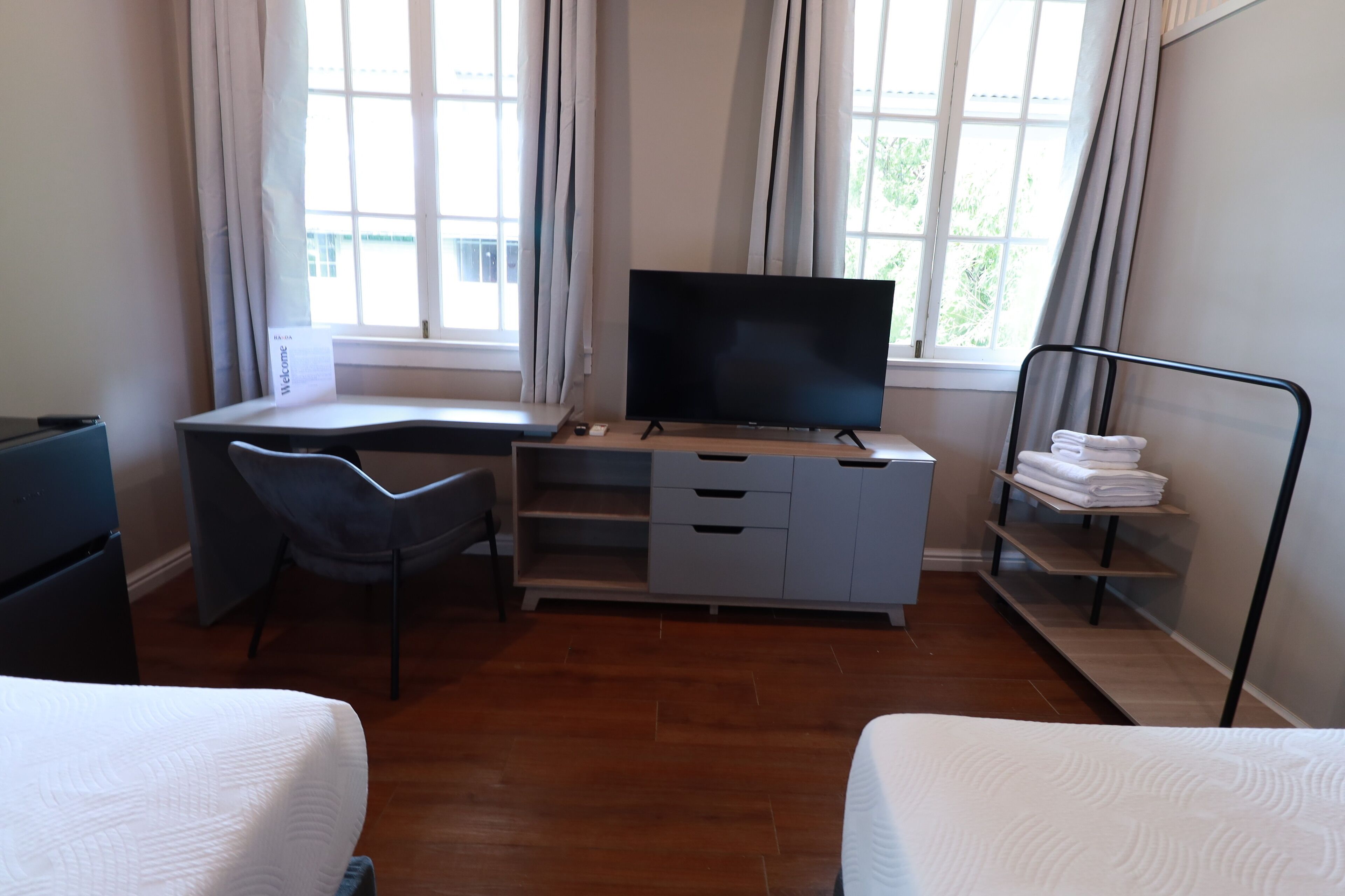 A room with a bed, a desk with a TV, a chair, and a shelf with towels.
