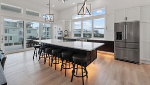 Cocina moderna con una isla grande, taburetes de bar, electrodomésticos de acero inoxidable y ventanas amplias que ofrecen vistas a casas vecinas.