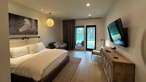 Ein modernes Hotelzimmer mit einem großen Bett, einem Flachbildfernseher, einer Holzkommode und Blick auf einen Garten durch eine Schiebetür.