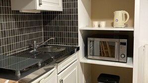 Una cocina compacta con salpicadero de azulejos negros, muebles de cocina blancos, un lavamanos de acero inoxidable, un microondas, una tetera y un cubo de basura.