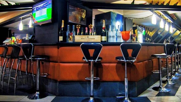A bar area with a checkered floor, a counter with bottles, and a TV screen. - Laroy Suites (Abeokuta)