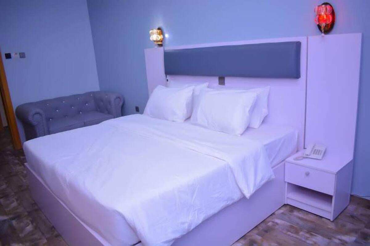 A bedroom with a large bed, a nightstand, a lamp, and a wall-mounted light fixture.