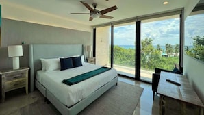 Une chambre à coucher moderne avec un grand lit, un ventilateur de plafond, une table de chevet avec une lampe et une vue sur l’extérieur grâce à de grandes fenêtres.