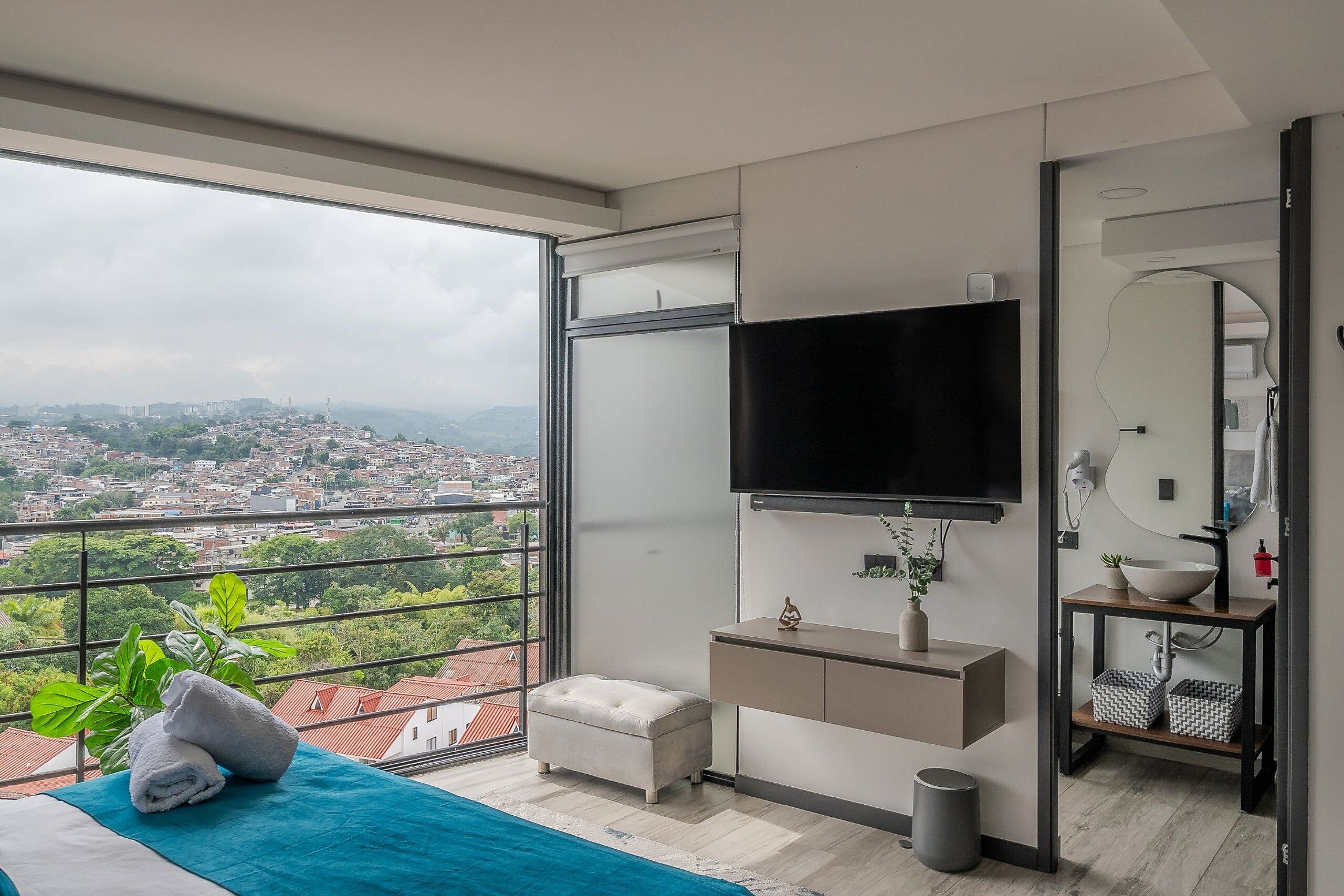 Ein modernes Schlafzimmer mit großem Fenster, Flachbildfernseher, Nachttisch, Spiegel und Blick auf die Stadt.